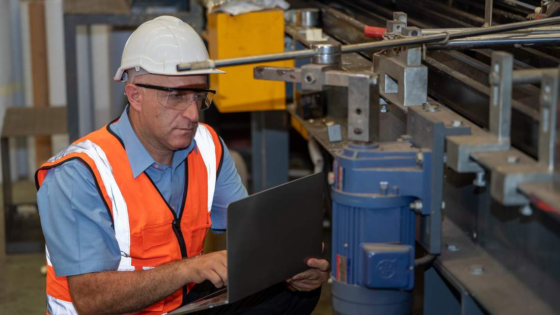 Plant engineer in a hard hat and safety vest using a laptop beside production machinery